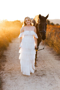 High school senior girl walking beside a horse on a dirt trail during golden hour, outdoor senior portrait by Kaitlyn Dawn Photography.