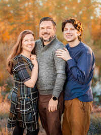 Family of three smiling together by a lake at golden hour during a New Bern family photography session