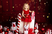 A young blonde girl in a red dress sits on a giant gift box surrounded by red and white Christmas decorations and a giant candy cane.