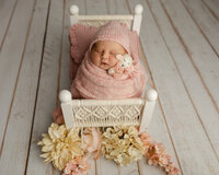 Sleeping newborn baby girl wrapped in a soft pink swaddle and posed in a miniature white bed prop during a professional studio newborn photography session.