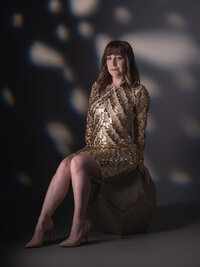 Woman in a gold sequined dress seated on a geometric block, dark studio background scattered with warm points of light.