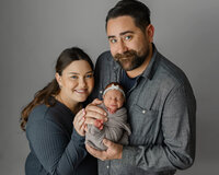 Parents smiling while holding their newborn baby wrapped in a soft gray swaddle during a studio newborn family portrait by Kaitlyn Dawn Photography.