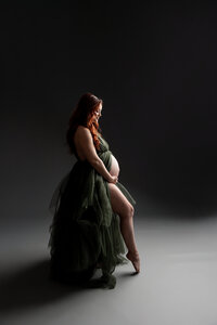 Fine art maternity portrait of a pregnant woman in a dark green gown standing in profile against a dramatic studio background by Kaitlyn Dawn Photography.