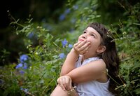 Jacksonville, FL portrait session capturing a Young girl sitting in a garden surrounded by greenery and blue flowers, gazing upward with a thoughtful look during an outdoor child portrait session in St. Johns, photographed by Nature's Child Photography