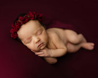 Newborn baby girl peacefully sleeping with hands under chin while wearing a burgundy headband on a red backdrop during a newborn photo session in Yucaipa by Kaitlyn Dawn Photography.