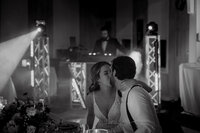 Mariés partageant un baiser tendre à leur table de réception, sous les lumières de la soirée, DJ en arrière-plan — instant authentique capturé par un photographe de mariage à Montréal, illustrant l’émotion et l’atmosphère festive de la célébration.