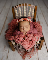 Sleeping newborn baby girl wearing a floral headband and posed in a small wooden chair with a textured pink blanket during a newborn photography session in Yucaipa by Kaitlyn Dawn Photography.