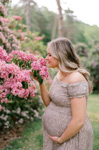 An expecting mom holding her belly while smelling a bush of pink flowers
