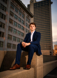 Senior portrait in downtown Mobile, Alabama— wearing a blue suit and brown dress shoes holding his hands together, buildings in background.