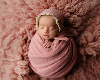 Sleeping newborn baby girl wrapped in a dusty rose swaddle and bonnet on a fluffy pink blanket during a studio newborn photography session by Kaitlyn Dawn Photography in Yucaipa.