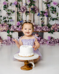 Smiling baby in a lavender “one” dress during a cake smash with purple flowers in the background. Photograph by Yucaipa cake smash photographer Kaitlyn Dawn Photography.