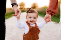 A toddler girl smiles while holding her parents hands.
