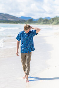 senior boy walking along a sandy beach and messing with his hair.