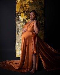A mother-to-be in an elegant copper dress stands confidently in front of a gold abstract backdrop, her gaze serene and poised.