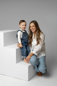 Mother sitting beside her toddler son on modern white studio blocks against a soft gray backdrop, smiling during a timeless studio family portrait session by Kaitlyn Dawn Photography.
