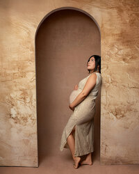 Expecting mom standing in an arched backdrop wearing a beige dress, highlighting her pregnancy during a maternity session by Kaitlyn Dawn Photography in the Inland Empire.