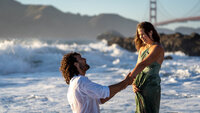 bearded man in a wet shirt on one knee proposing lovingly to a beautiful woman holding the edge of her dress from the water with the surf and golden gate bridge in the background.