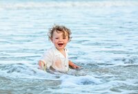 Ponte Vedra portrait session capturing Laughing toddler boy in a white button-down shirt sitting in shallow ocean waves as water splashes around him during a beach child portrait session photographed by Nature's Child Photography