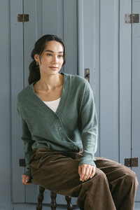 Model in a sage green V-neck cardigan and brown corduroy trousers seated against a painted boathouse door