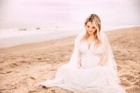 A pregnant woman with blonde hair sits on the beach in a white dress. The shoreline runs diagonally to her and the sky is subtly pink.