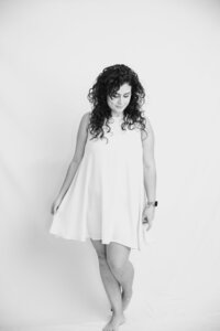 A black and white picture of a woman with dark curly hair smiling softly down, standing on a white backdrop. She wears a white dress and holds it loosely away from her body.