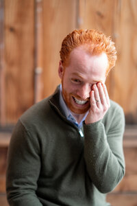 Bearded man wearing a sweater in an office laughing while holding his face