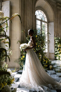 Bride in a strapless lace gown holding white flowers, standing in a sunlit hall with tall windows, lush greenery, and floral arrangements on a checkered floor.