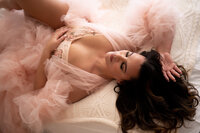 A brunette woman dressed in a fluffy pink robe and pink floral lingerie reclines for a studio portrait.
