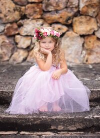 Jacksonville, FL portrait session capturing a thoughtful young princess in a pastel tulle dress and floral crown as she sits on rustic stone steps during a signature fairy adventure session in Palatka.