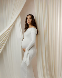Pregnant woman in an elegant white dress posing beside soft neutral drapery, showcasing her pregnancy during a Yucaipa maternity session with Kaitlyn Dawn Photography.