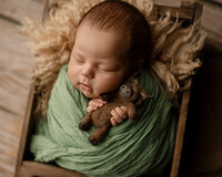 Sleeping newborn baby boy wrapped in a green swaddle and holding a small teddy bear while posed in a wooden crate during a studio newborn photography session by Inland Empire newborn photographer Kaitlyn Dawn Photography.