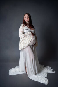 A pregnant woman in a long, lace-sleeved white gown poses against a dark gray backdrop, exuding elegance and grace.