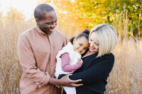 A man and woman stand in a field of tall grass, smiling and embracing a young girl between them, with autumn trees in the background—a perfect moment captured by a Toronto Family Photographer.