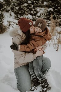 A mother snuggles her young son in the snow during a New England winter. Fine art lifestyle family photography by Damini Photography capturing warmth in the winter.