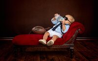 Baby boy wearing sunglasses and suspenders relaxes on a red chaise with a football beside him while drinking from a bottle during a studio child portrait session in Mandarin. - A portrait experience by Nature's Child Photography.