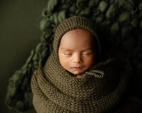 Sleeping newborn baby wrapped in a cozy olive knit swaddle and bonnet on a dark green textured backdrop during a studio newborn photography session by Kaitlyn Dawn Photography.