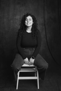A black and white picture of a woman with dark curly hair sitting on a white stool against a dark gray backdrop. She wears jeans and a black long-sleeve shirt. holds the stool between her legs and smiles ahead.