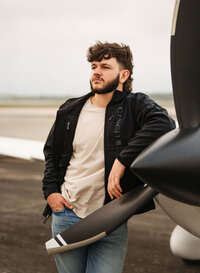 Senior portrait at a small hangar near Mobile International Airport in Mobile, Alabama — leaning
against the propeller in a black jacket, looking off into the distance.