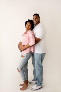 smiling expecting couple standing while holding baby bump in studio portrait for maternity photoshoot in North Hollywood