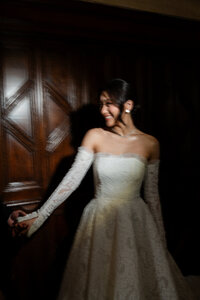 Bride in a high-fashion, off-the-shoulder lace gown with long gloves, featuring a polished Kohnur elegant bridal updo and flawless timeless wedding makeup, posing in a dramatic, dimly lit setting.