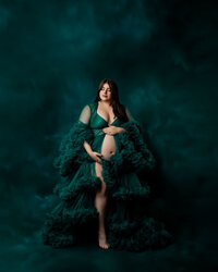 Expecting mother posing in an emerald green ruffled maternity gown against a dark studio backdrop during an artistic maternity photography session by Kaitlyn Dawn Photography.