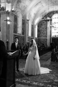 A bride and groom stand holding hands before a minister in an ornate church, with stone walls and a high, decorated ceiling. The scene is black and white, and guests are seated in pews, observing the ceremony.