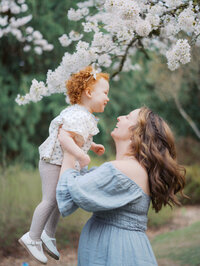 Mom is laughing and lifting her red-haired toddler up in the air