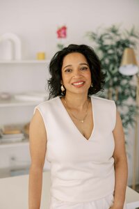 Woman leaning casually on a table, wearing an all white outfit and smiling at the camera