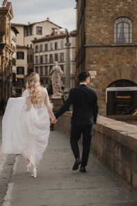 Mariés courant main dans la main dans une rue pavée historique, robe vaporeuse à manches longues et complet noir, statue et architecture italienne en arrière-plan, mariage à destination en Italie