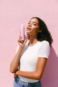 Model in a white tee against a pink wall holding Evian Glow facial mist