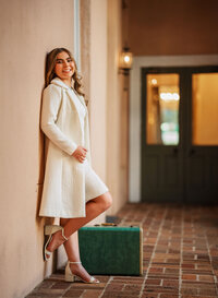 Senior portrait at the Malaga Inn in Mobile, Alabama— wearing a vintage white coat and dress leaning against a while stand next to a green suitcase.