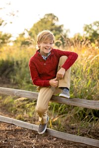 Carefree tween boy in O’Fallon, Illinois smiles confidently while perched on a rustic wooden fence, golden sunset light glowing through tall autumn grasses during an outdoor. (Photographed by Nature's Child Photography)