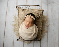 Sleeping newborn baby girl wrapped in a cream swaddle and posed in a vintage baby bed prop on a neutral wood backdrop during a newborn photography session by Kaitlyn Dawn Photography in Yucaipa.