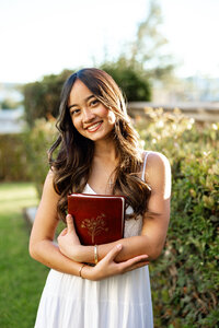 High school senior girl holding a book and smiling in a garden setting, outdoor senior portrait by Kaitlyn Dawn Photography.
